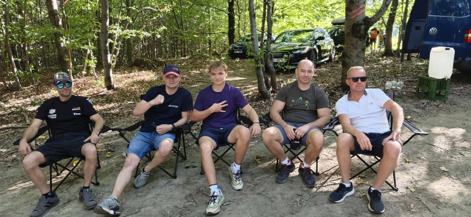 Pięciu uśmiechniętych mężczyzn siedzi na krzesłach turystycznych w lesie na tle zaparkowanych samochodów podczas rajdu.